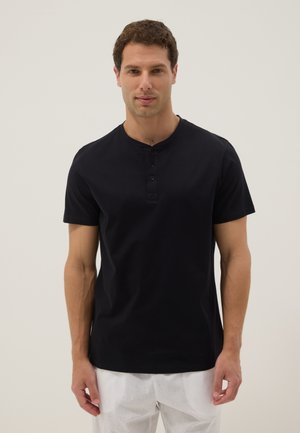 Homme portant une chemise henley noire à manches courtes et un pantalon blanc, debout devant un fond clair uni, faisant face à l’avant.