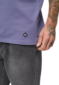 Lilla bomulds t-shirt med korte ærmer, prydet med et lille sort diamanformet logo ved kanten, båret med grå denim jeans.