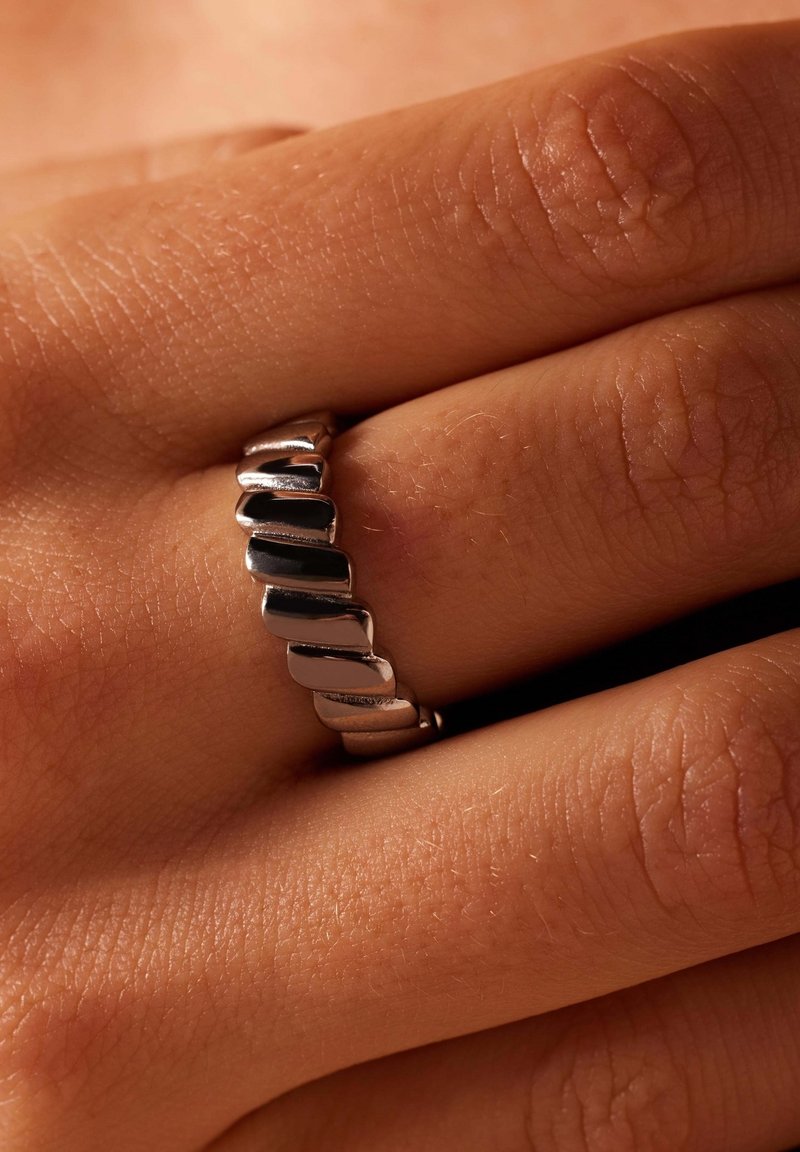 Zilveren ring met een golvend ontwerp, bestaande uit gepolijste en matte secties, geplaatst op een gestructureerde huidachtergrond met twee zichtbare vingers.