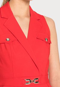 Sleeveless red blazer dress with lapel collar, two flap pockets with gold buttons, and a matching belt with a gold buckle.