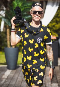 Schwarzes Shirt und Shorts mit gelben Gummi-Entenmuster. Das Modell hält eine Kamera und trägt Sonnenbrillen, mit einem sichtbaren Tattoo auf einem Arm.