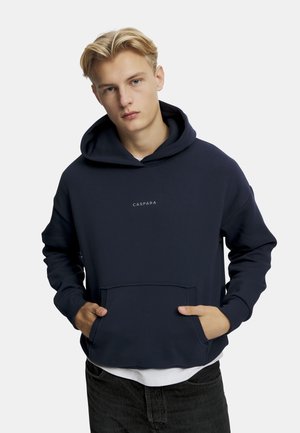 Marinblå hoodie i mjukt tyg, med en känguruficka, dragsko i huvan och en liten logga på bröstet. Bärs över en vit skjorta.