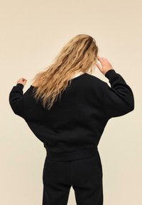 Crna široka majica s velikim okovratnikom i spuštenim ramenima, izrađena od teksturiranog materijala s rebrastim manšetama, u kombinaciji s pripadajućim hlačama.