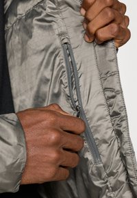 Könnyű szürke dzseki belseje sima textúrájú; egy cipzáras zsebhez való hozzáférés látható egy kéz által, bemutatva a cipzár fémalkatrészeit.