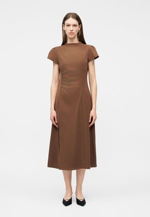 Moteris stovinti tiesiai, dėvinti rudonos spalvos midi suknelę trumpomis rankovėmis ir juodus smailianosčius batelius, priešais paprastą baltą foną.