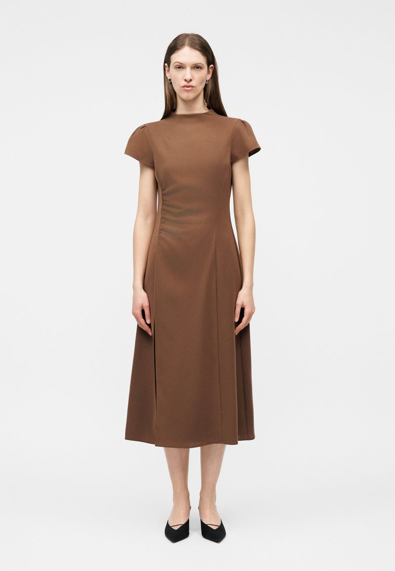 Γυναίκα που στέκεται όρθια, φορώντας ένα καφέ μίντι φόρεμα με κοντά μανίκια και μαύρα παπούτσια με μυτερή μύτη, σε απλό λευκό φόντο.