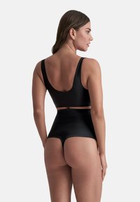 Kvinna som bär svarta högmidjade stringtrosor och en svart ärmlös crop top, sedd bakifrån mot en enfärgad bakgrund.