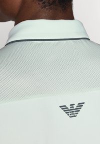 Φωτεινό πράσινο, υφασμάτινο πόλο μπλουζάκι με διάτρητο σχέδιο και σκούρο γκρι περιθώριο. Διαθέτει μαύρο λογότυπο στην πλάτη.