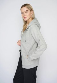 Siva zip-up dukserica napravljena od mekog materijala, s kapuljačom, prednjim džepovima i podesivim špagom na rubu. Jednostavan, ležeran dizajn.