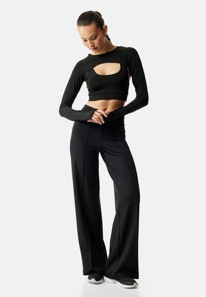 Mujer con crop top negro de manga larga con abertura en el pecho, pantalones negros de pierna ancha y cintura alta, y zapatillas negras, posando con las manos cruzadas.