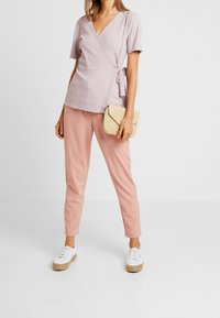 Top envolvente de color púrpura claro con mangas cortas, combinado con pantalones ceñidos rosas y zapatillas blancas. Se incluye un pequeño bolso cruzado de tejido.