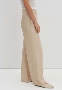 Pantalons larges beiges en tissu lisse, avec des poches latérales et une taille épurée. Associés à des chaussures plates de couleur claire.