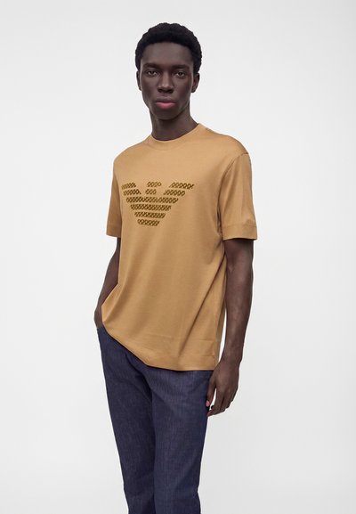 Άνδρας που φοράει καφέ κοντομάνικο πουκάμισο με σκούρο λογότυπο με μοτίβο και σκούρο μπλε παντελόνι, στέκεται μπροστά σε ένα απλό φωτεινό φόντο.