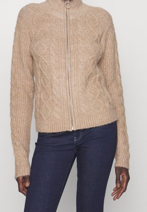 Beige kabelgebreide trui met rits en hoge kraag, gedragen met donkerblauwe aansluitende jeans, getoond van nek tot heupen.