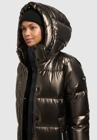 Fémes bronz színű puffadt dzseki nagy kapucnival, fekete cipzárral és patent gombokkal. Bal ujján logófolttal.