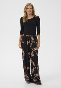 Glunderende vrouw met lang blond haar, gekleed in een zwart langemouwen top en zwarte wijde broek met een bruine paisley print, staande op een witte ondergrond.