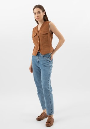 Žena stojí s pravou rukou v zadní kapse, má na sobě bezrukávou hnědou vestu na knoflíky, světle modré džíny a hnědé tenisky na bílém pozadí.