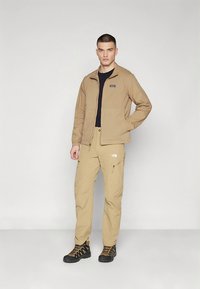 Jachetă bej cu fermoar în față, cămașă neagră dedesubt, pantaloni bej cu buzunare și bocanci de drumeție. Jacheta are un logo pe piept.