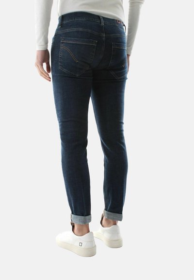 Jeans in denim blu scuro con taglio slim, dotati di orli arrotolati, tasche posteriori e dettagli di cucitura sottili. Abbinati a sneakers bianche.