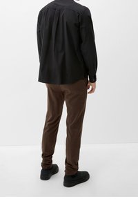 Camisa negra de manga larga con un pliegue en la parte trasera, combinada con pantalones marrones texturizados y zapatos negros, vista desde atrás sobre un fondo blanco.