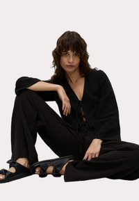 Czarna, lekka bluzka z długimi rękawami i detalem w postaci kokardy, zestawiona z czarnymi, szerokimi spodniami oraz płaskimi sandałami z akcentami w postaci kokard.