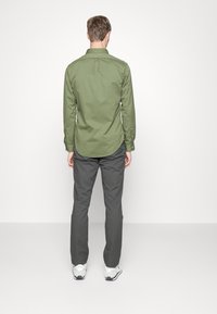 Grünes Button-Down-Hemd mit langen Ärmeln und abgerundetem Saum, kombiniert mit grauen Hosen und weißen Sportschuhen. Von hinten gezeigt.