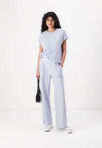 Jumpsuit a righe blu e bianche con maniche corte, dettaglio anteriore annodato, gambe ampie e dettaglio con bottone. Inclusi borsa nera e sneakers.