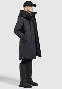 Black insulated parka featuring a hood, two side pockets, and a button closure. Worn with black leggings and knee-high rubber boots.