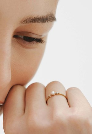 Primer plano de un ojo cerrado y una mano con un anillo de oro que tiene una pequeña perla y dos diminutos diamantes en el dedo anular.