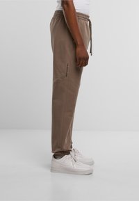 Bruine sweatpants met een elastische tailleband en trekkoord, met zijnaad en een logo op het linkerbeen gedrukt. Draagt een paar witte sneakers.
