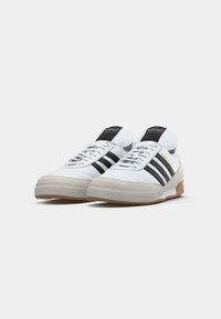 Białe tenisówki Adidas z czarnymi paskami, beżowymi zamszowymi czubkami i gumowymi podeszwami, ustawione obok siebie na białym tle.