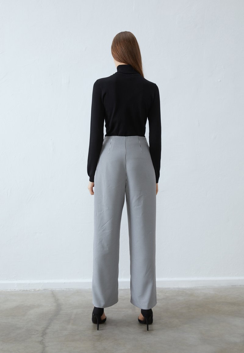 Maglione nero a collo alto con maniche aderenti abbinato a pantaloni a vita alta e gamba larga in un motivo a quadri bianco e nero. Scarpe con tacco.