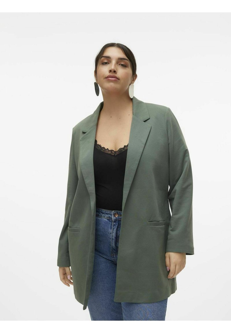 Veste olive verte avec une coupe décontractée, des manches longues et deux poches avant, accompagnée d'un haut noir avec des bordures en dentelle et d'un jean en denim bleu.