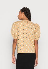 Blusa de mangas curtas em mostarda com estampa de flores brancas, apresenta mangas bufantes com franzido e uma abertura em forma de lágrima nas costas. Textura de algodão suave.