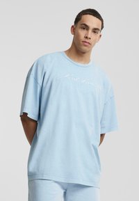 T-shirt surdimensionné bleu clair en coton, avec un col rond et des manches courtes, arborant un texte brodé blanc à l'avant.