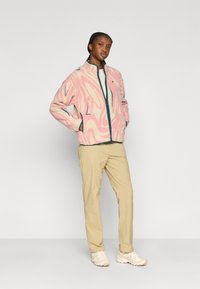 Φλις μπουφάν σε ροζ με κυματιστά σχέδια, ναυτικά μπλε στοιχεία, όρθιο φερμουάρ και εμπρόσθιες τσέπες. Μπεζ παντελόνια και ελαφριά αθλητικά παπούτσια ολοκληρώνουν την εμφάνιση.