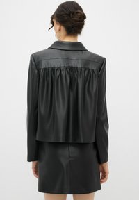 Schwarze Kunstlederjacke mit gerafftem Rückendetail, spitzem Kragen und langen Ärmeln, kombiniert mit einem passenden Minirock.