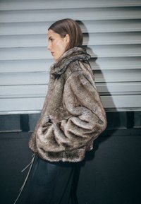 Barna műszőrme dzseki magas gallérral, széles ujjakkal és elülső zsebekkel, fekete mintás nadrág felett viselve, szürke háttér előtt.