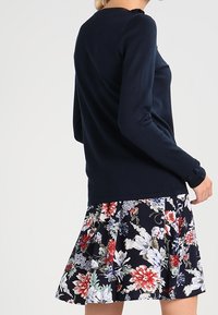 Marinblå långärmad topp i kombination med en blommönstrad kjol med röda, vita och blå blommor mot en mörk bakgrund. Mjuk kostym, figurnära design.