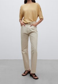 Kvinna iklädd en beige kortärmad topp, ljusbeige högmidjade byxor med bälte och svarta remsandaler med klack, ståendes med händerna i fickorna.