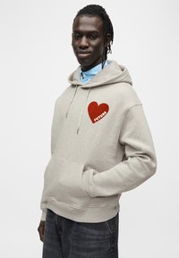 Sweat à capuche gris avec une poche avant, arborant un graphique de cœur rouge et le mot "FUTURE". Porté sur une chemise à col bleu clair.
