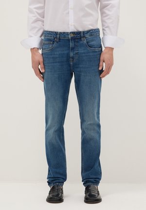 Uomo che indossa jeans blu a gamba dritta, mocassini neri e camicia bianca a maniche lunghe con polsini arrotolati.