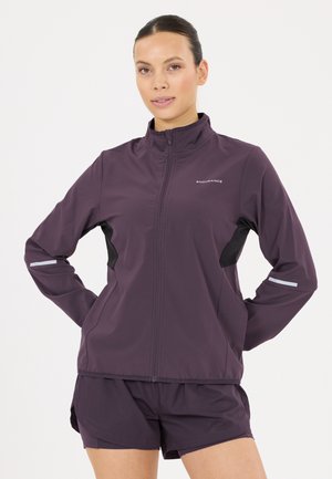 Vrouw die een paarse sportjas met rits en bijpassende korte broek draagt, staat met haar handen achter haar rug tegen een effen achtergrond.