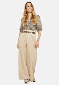 Bēša plaša stila bikšu ar locījumiem un brūnu jostu, kopā ar īsroku leoparda raksta blūzi un rakstainām sporta kurpēm.