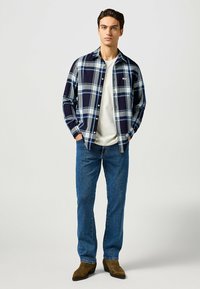 Camicia di flanella a quadri nei colori navy, grigio e azzurro, con chiusura a bottoni, indossata su una maglietta bianca con jeans blu e stivali marroni.