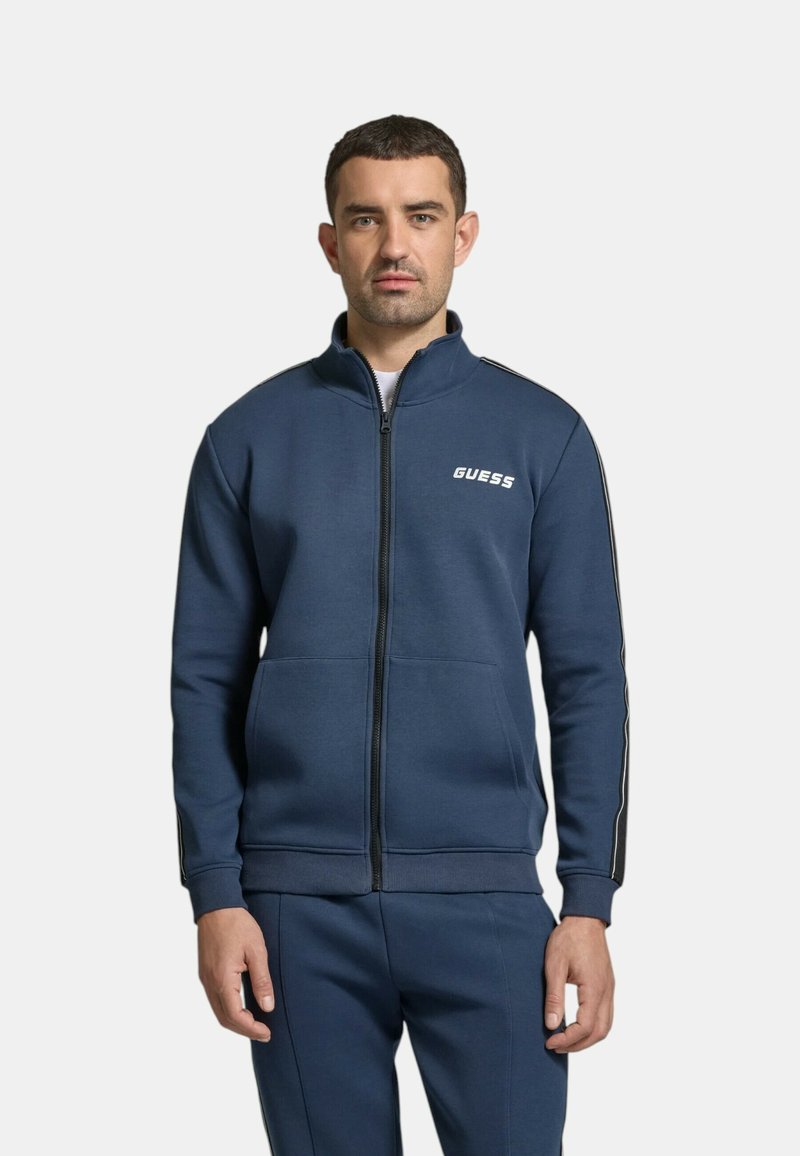 Felpa blu navy con zip, colletto alto, polsini a coste e tasche. Presenta un logo bianco e strisce laterali nere a contrasto. Morbido tessuto in misto cotone.