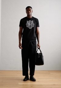 Zwarte T-shirt met een zilveren leeuwengrafiek, zwarte broek en zwarte leren tas. De achtergrond is een lichtgrijze muur met een houten vloer.