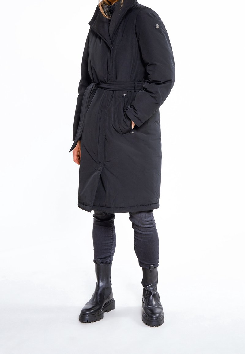Sort lang dunjakke med bælte i taljen, høj krave og sidelommer. Båret over mørke jeans og sorte kraftige støvler. Glat tekstur.