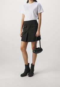 Mujer vistiendo una camiseta blanca, falda negra con bolsillos frontales, botas negras hasta el tobillo, sosteniendo un pequeño bolso negro frente a un fondo simple.