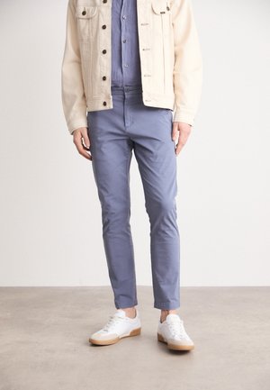 Uomo con giacca beige chiaro, camicia blu, pantaloni blu e sneakers bianche in piedi su pavimento grigio davanti a uno sfondo bianco.